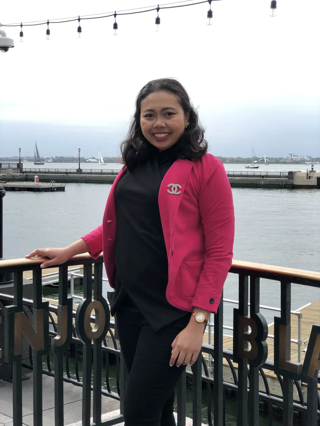 Krittaya leaning on a railing by the water, wearing a pink blazer